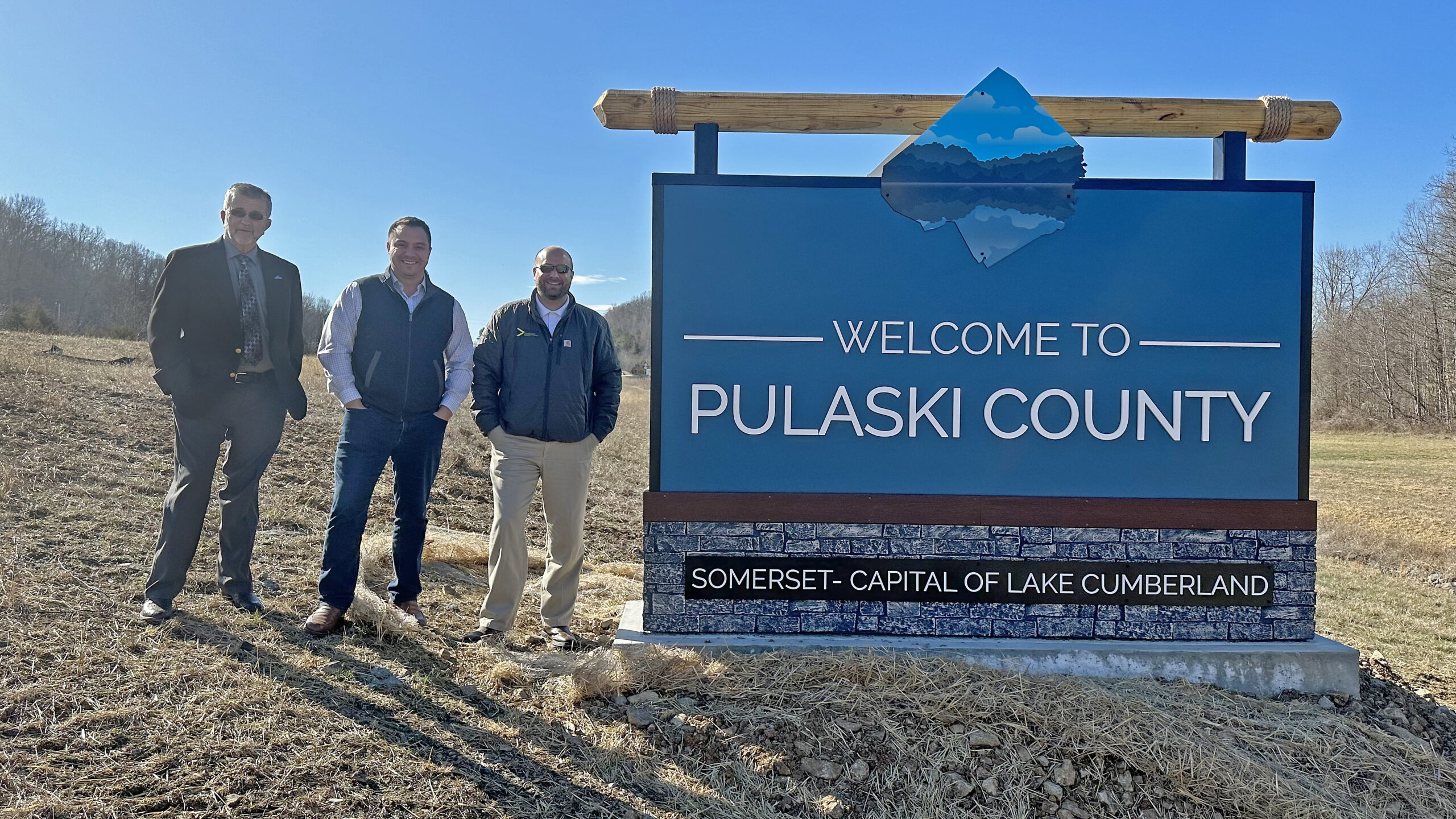 Three men standing to the left of a sign that says Welcome to Pulaski County