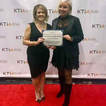 two women in formal wear standing on red carpet in front of backdrop holding certificate