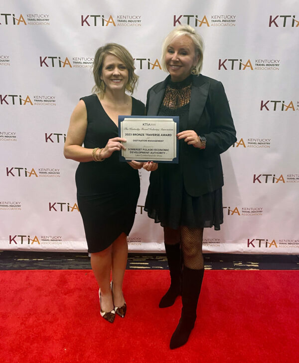 two women in formal wear standing on red carpet in front of backdrop holding certificate