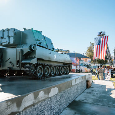 military artillery vehicle on top of a concrete stage with an American flag high on a fire truck in the background